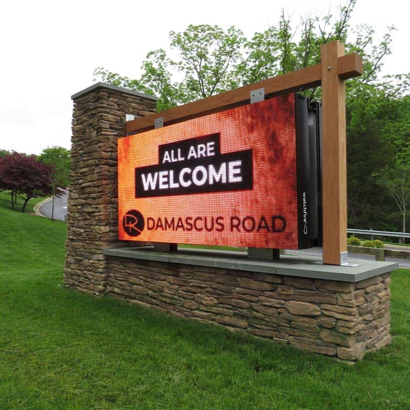 Digital LED monument sign with stone brick base and mounted wood beams at the entrance of Damascus Road Community Church