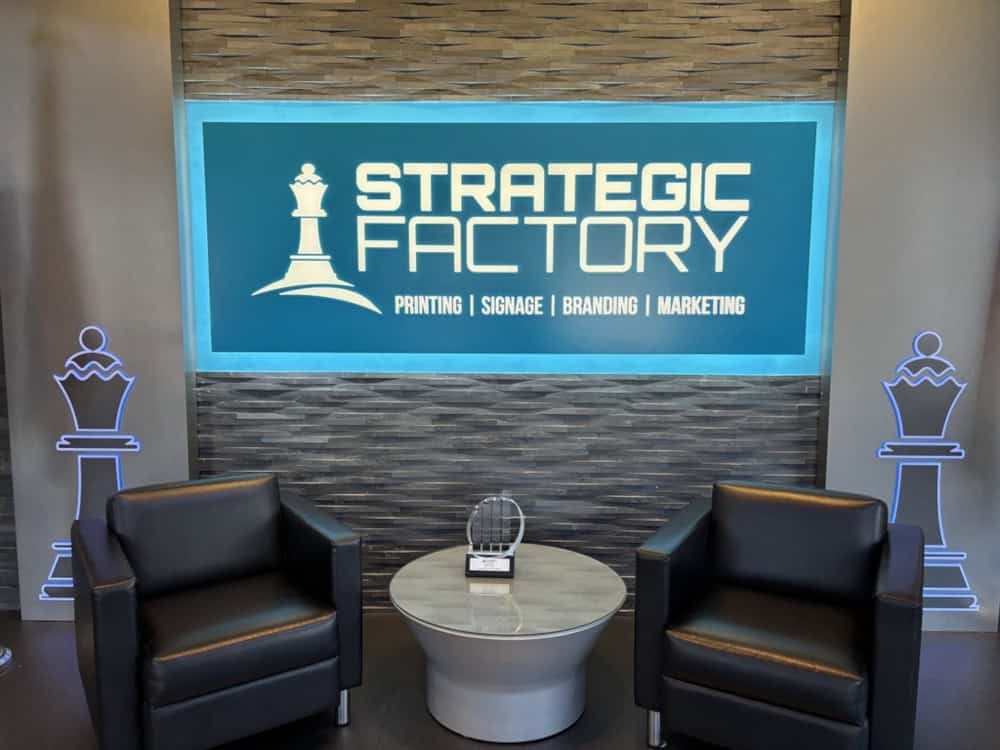Strategic Factory lobby with a backlit company sign displaying 'Printing | Signage | Branding | Marketing,' flanked by two black chairs and a round table with an award.